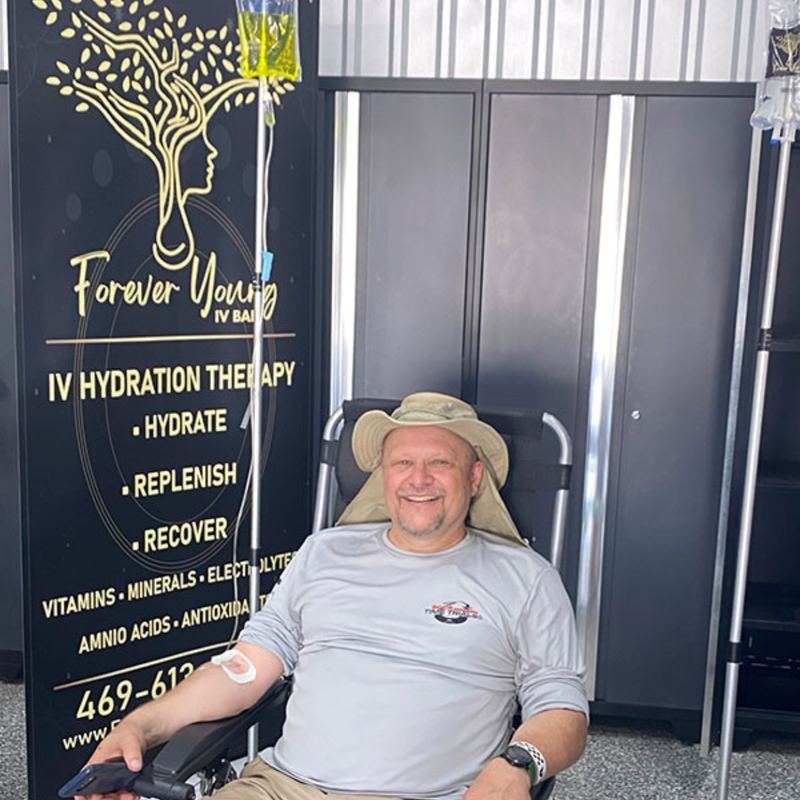 A client on an IV drip beside the Forever Young IV Bar banner at an event
